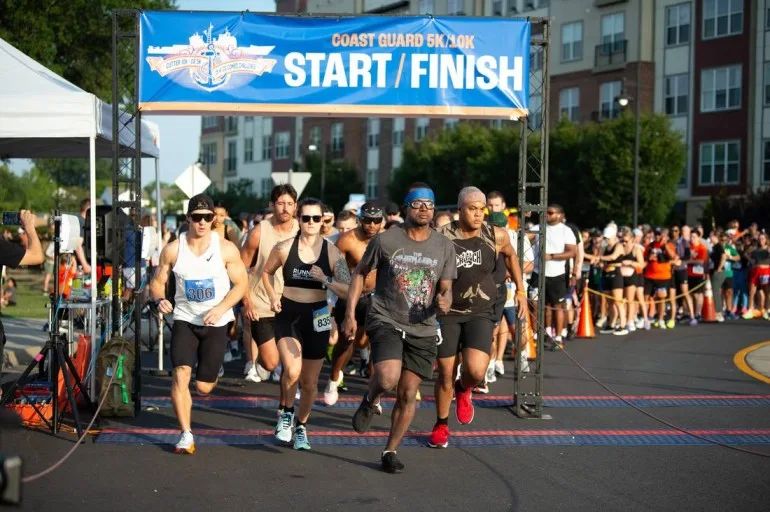 Runners bursting across the mat under the Coast Guard 5K/10K start and finish banner in Portsmouth